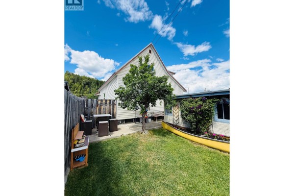 Backyard view of a white house with green grass, a wooden fence, patio furniture, and a yellow canoe planter. - 266 Ritchie Street, Kimberley