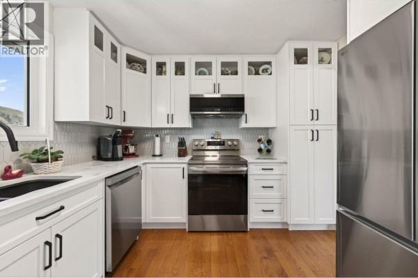 Modern white kitchen with stainless steel appliances, quartz countertops, and a breakfast bar with stools. - 2660 Argyle Avenue, Kamloops