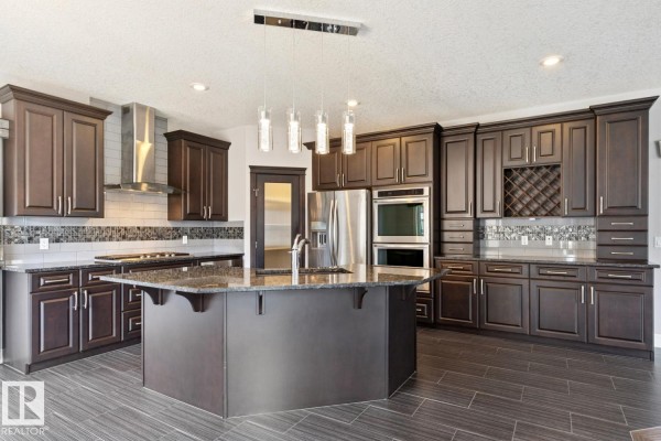 Kitchen with dark wood cabinets, granite countertops, stainless steel appliances, and a large island with a sink. - 2665 Watcher Wy Sw, Edmonton