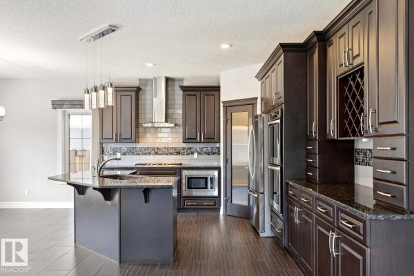 Kitchen area with dark wood cabinetry, granite countertops, stainless steel appliances, and a view into a dining space. - 2665 Watcher Wy Sw, Edmonton