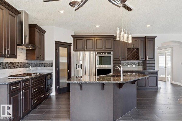 Spacious kitchen featuring dark wood cabinets, a large island, stainless steel appliances, and a wine rack display. - 2665 Watcher Wy Sw, Edmonton