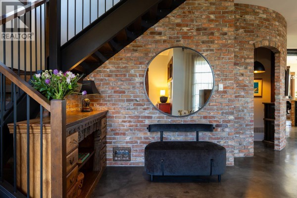 Interior entryway with a brick accent wall, a metal staircase, a wooden console, and a round mirror. - 2673 Bath Street, Kelowna