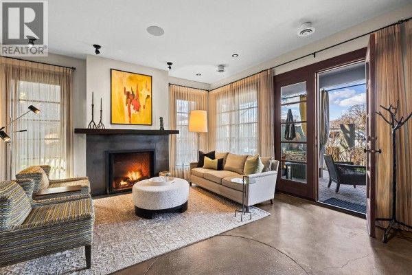 Living room with a fireplace, sofa, two armchairs, and large glass doors opening to an outdoor patio. - 2673 Bath Street, Kelowna