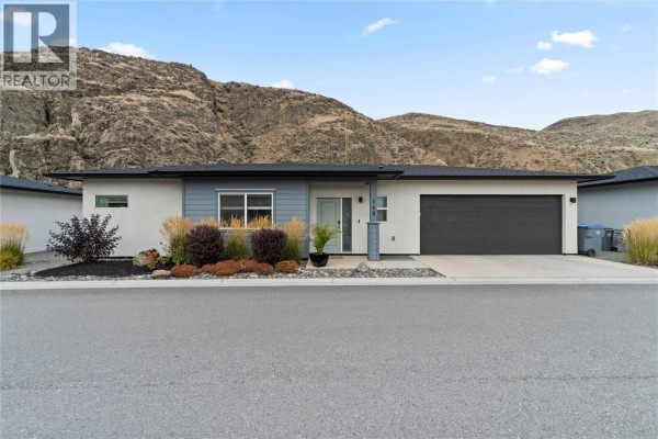 Modern single-story home with a dark grey garage door and mountain backdrop during the day. - 2683 Ord Road Unit# 148, Kamloops