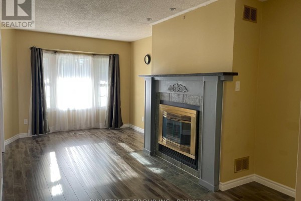 Living room with bay windows, dark curtains, wood-look flooring, and a grey fireplace with a brass insert. - 27 Lindsay Court, Barrie