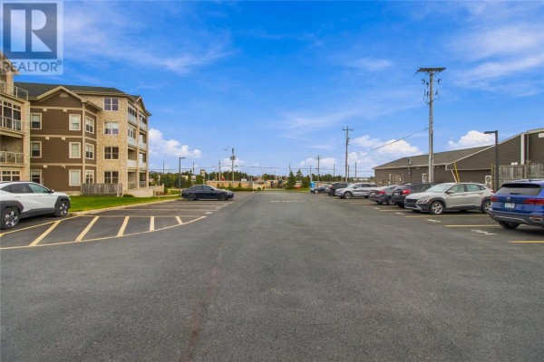 View down a parking lot with cars. The apartment building's tan exterior is on the left, and other buildings are on the right. - 27 Rhodora Street Unit#306, St  John S