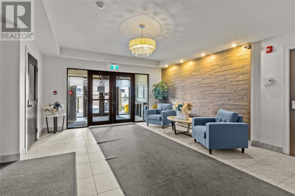 Building lobby with tiled floors, gray carpet runner, and two blue armchairs. A stone accent wall is behind the seating. - 27 Rhodora Street Unit#306, St  John S
