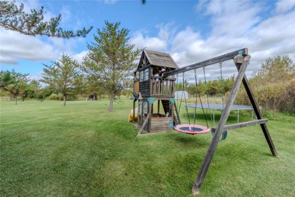 Wooden play structure with a swing set, slide, and trampoline on a green grassy lawn. - 270 Seine River Trail, La Broquerie