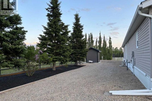 Side view of the mobile home with a gravel driveway, shed, and mature trees. - 270165 Range Road 291  Se, Airdrie