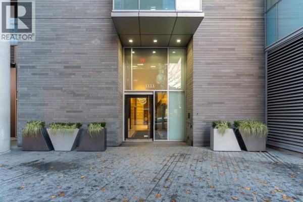 Building entrance with glass doors, grey brick facade, and modern planters with greenery. - 2704 1111 Alberni Street, Vancouver