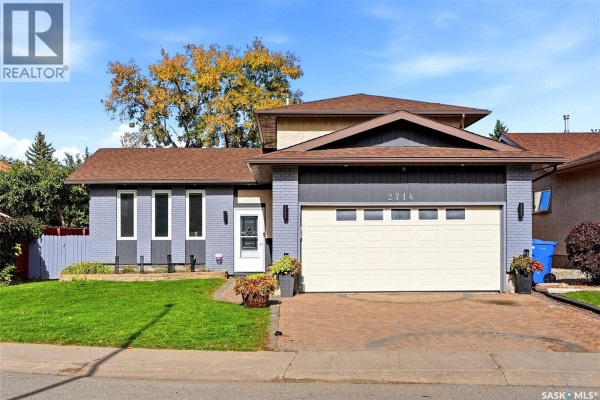 Front exterior view of a residential house with a beige and grey facade, garage door and lawn. - 2714 Jaques Place E, Regina