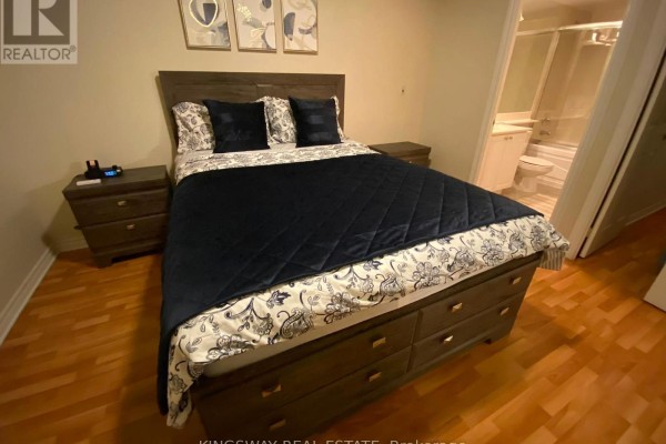 Bedroom with queen-size bed, navy bedding, wood nightstands, and view into a bathroom. - 2730 - 3888 Duke Of York Boulevard, Mississauga