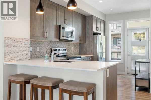 Kitchen island with white quartz countertop, stainless steel dishwasher, and three upholstered stools. - 28, 138 Seton Passage Se, Calgary