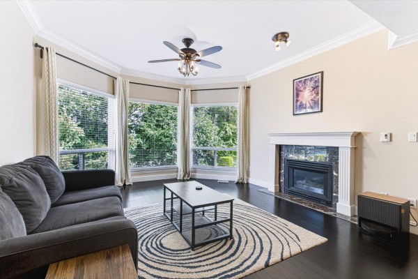 Living room fireplace with marble surround, grey sofa, and large windows viewing trees. - 28 32777 Chilcotin Drive, Abbotsford