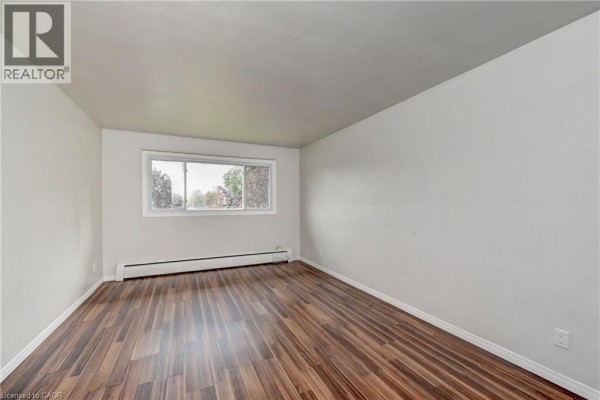 Living room with dark wood laminate flooring, white walls, and a window showing trees outside. - 280 Sydney Street S Unit# A6, Kitchener