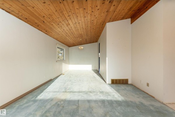Spacious room with a wood-plank ceiling, light blue carpet, and a small window showing a view of houses and trees. - 280 Warwick Rd Nw, Edmonton