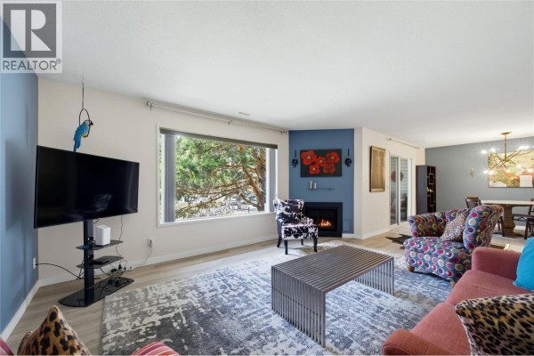 Living room with fireplace, TV, patterned chairs, and a window looking out to trees. - 2806 35 Street Unit# 207, Vernon