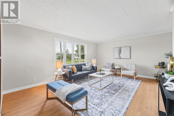 Living room with light grey walls, a large window, dark grey sofa, blue ottoman, and wooden floors. - 283 Fowlers Road, Conception Bay South