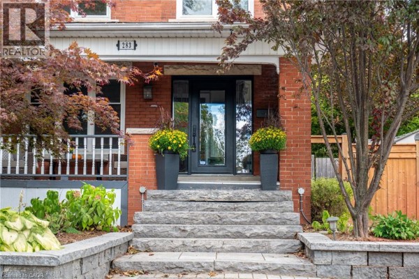Side view of the red brick house with a porch, driveway, and wooden fence. - 283 Plains Road W, Burlington