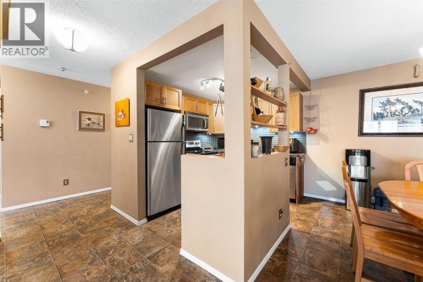 Dining area with wooden table and chairs, beige walls, and a mirror reflecting the kitchen. - 284 Yortkton Avenue Unit# 205, Penticton