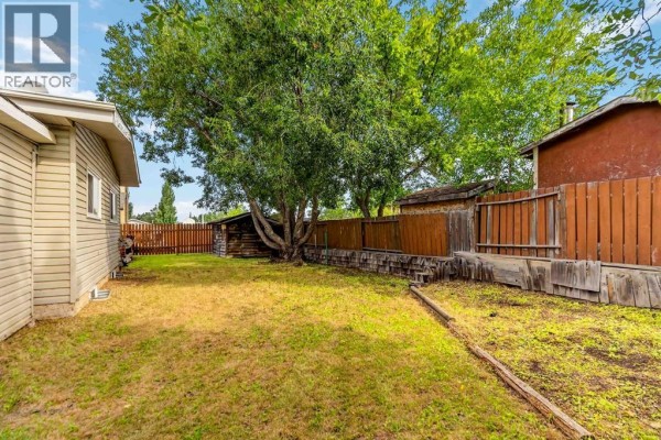 View of a backyard with green grass, wooden fence, and storage sheds under a large tree. - 286 Kennedy Bay, Fort Mcmurray