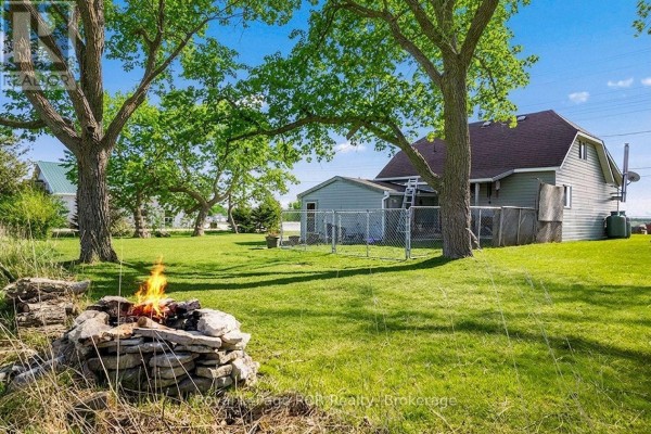 Rear view of the light gray house with a chain-link fence and a stone fire pit. - 2882 6 Highway, Northern Bruce Peninsula