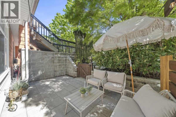 Outdoor patio with light-colored furniture, umbrella, and stone retaining wall surrounded by lush greenery. - 29 6878 Southpoint Drive, Burnaby