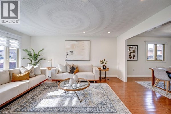 Living room with two beige sofas, a patterned area rug, wood floors, and large windows. - 29 Napoleon Drive, Stoney Creek