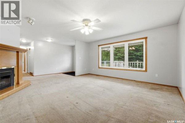 Living room with a beige carpet, large window overlooking trees, and a fireplace. - 292 Regina Avenue, Katepwa Beach