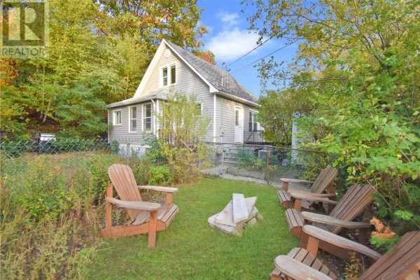 Backyard with Adirondack chairs arranged around a fire pit in green grass. - 294 Kathleen Street, Sudbury