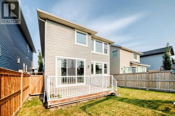 Backyard with a wooden deck with white railings and stairs, leading to a two-story grey house with sliding doors. - 296 Nolancrest Heights Nw, Calgary