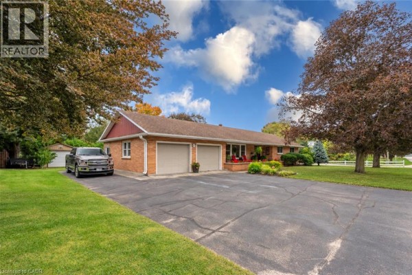 Driveway and side view of the brick house with a two-car garage, surrounded by green lawn and trees. - 297 Main Street W, Otterville