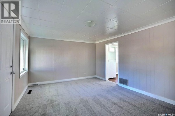 Empty living room with grey carpet, light tan paneled walls, white trim, and a doorway. - 297 Smith Street, Regina