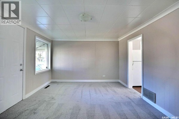 Living room showing a large window, grey carpet, paneled walls, white trim, and a doorway to the kitchen. - 297 Smith Street, Regina