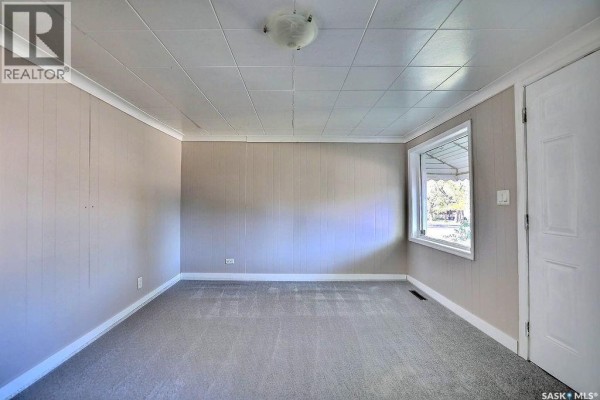 Empty room with grey carpet, paneled walls, white trim, a large bay window, and a white door. - 297 Smith Street, Regina