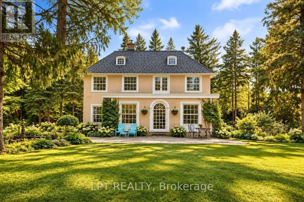 Front view of a light yellow stucco house with manicured gardens, a patio with seating, and bright blue Adirondack chairs on the lawn. - 298 Wees Road, Uxbridge