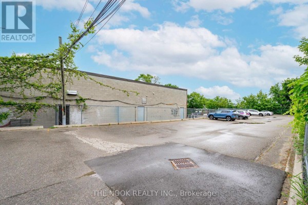 Rear asphalt parking lot with cars parked, bordered by a chain-link fence and overgrown green vines. - 2b - 337 King Street W, Oshawa