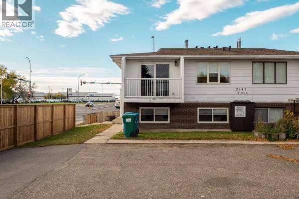Townhouse exterior, beige siding, white trim, brick base, and a wooden fence. Parking lot visible. - 3, 2145 5 Avenue N, Lethbridge