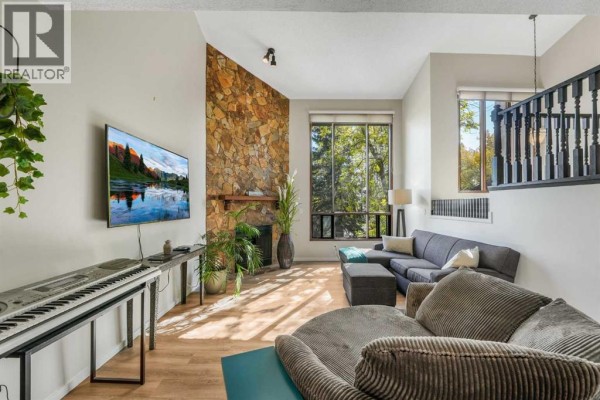 Living room with a stone fireplace, large windows, grey sectional sofa, and a keyboard. - 3, 7900 Silver Springs Road Nw, Calgary