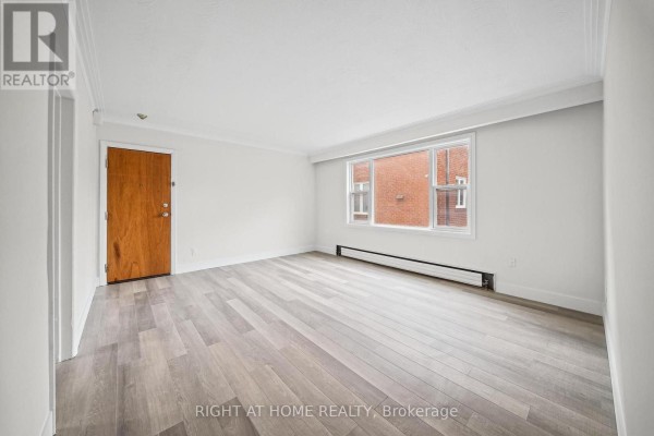 Empty living area with light wood floors, a large window facing a brick building, and a wooden door. - 3 - 32 Vanevery Street, Toronto