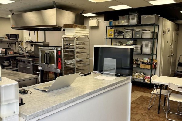 Commercial kitchen with stainless steel appliances, oven, shelves, and a counter with a laptop. - 3 - 5360 Canotek Road, Ottawa
