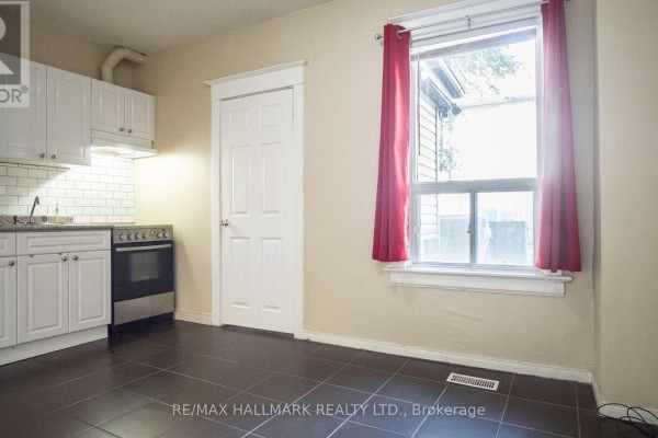 Kitchenette with white cabinets and appliances, a white door, and a window with red curtains. - 3 - 560 Jones Avenue, Toronto