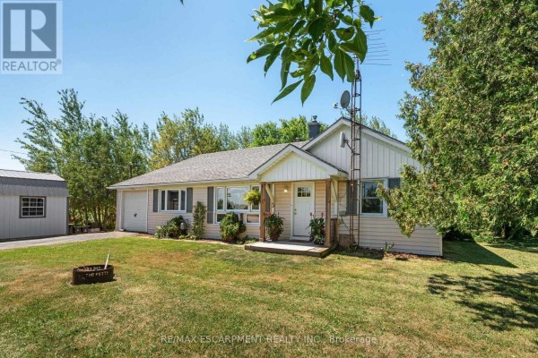 Side view of a beige bungalow with a porch, featuring wood columns, flowers, and a fire pit in the yard. - 3 Bousfield Lane, Haldimand