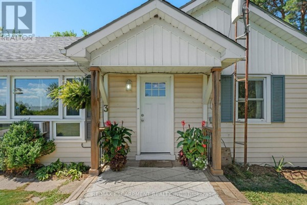 Close-up of the beige bungalow's covered front porch entrance with a white door, windows, and potted plants. - 3 Bousfield Lane, Haldimand