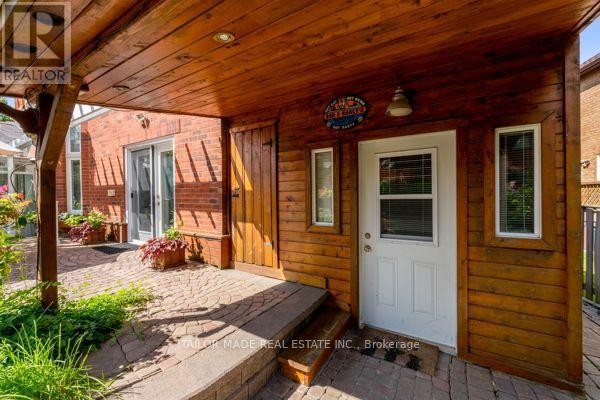 Covered wooden porch area with a brick patio, white door, and adjacent brick wall. - 3 Stonegate Drive, Brampton