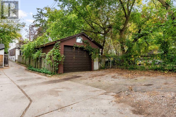 Rear view of a detached wooden garage with a brown door and overgrown greenery in a gravel yard. - 30 Lawlor Avenue, Toronto