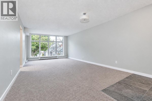 Empty living room with large windows, grey carpet, and tiled accent floor. - 300 2533 Penticton Street, Vancouver