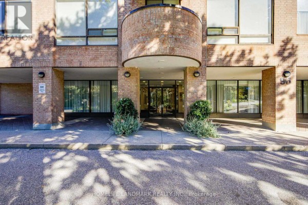 Entrance to a brick apartment building with glass doors, flanked by bushes and a paved walkway. - 301 - 96 Fifeshire Road, Toronto