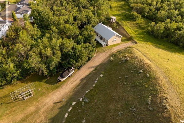 Aerial view of the house situated on a hill, surrounded by trees, with a boat and trailer below. - 301 Pine Tree Road, Buffalo Pound Lake