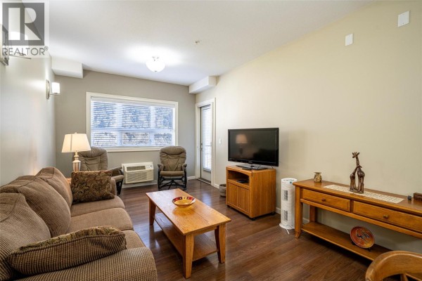 Living room with a brown sofa, coffee table, television, and window with blinds. - 3010 35 Street Unit# 207, Vernon